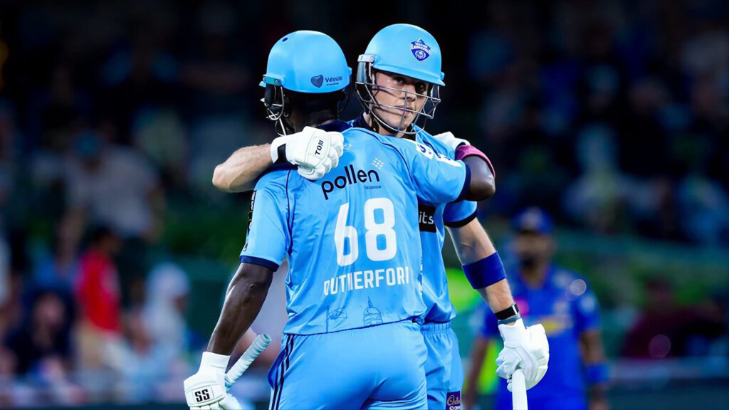 Two cricket players in blue uniforms embrace on the field after a successful play.